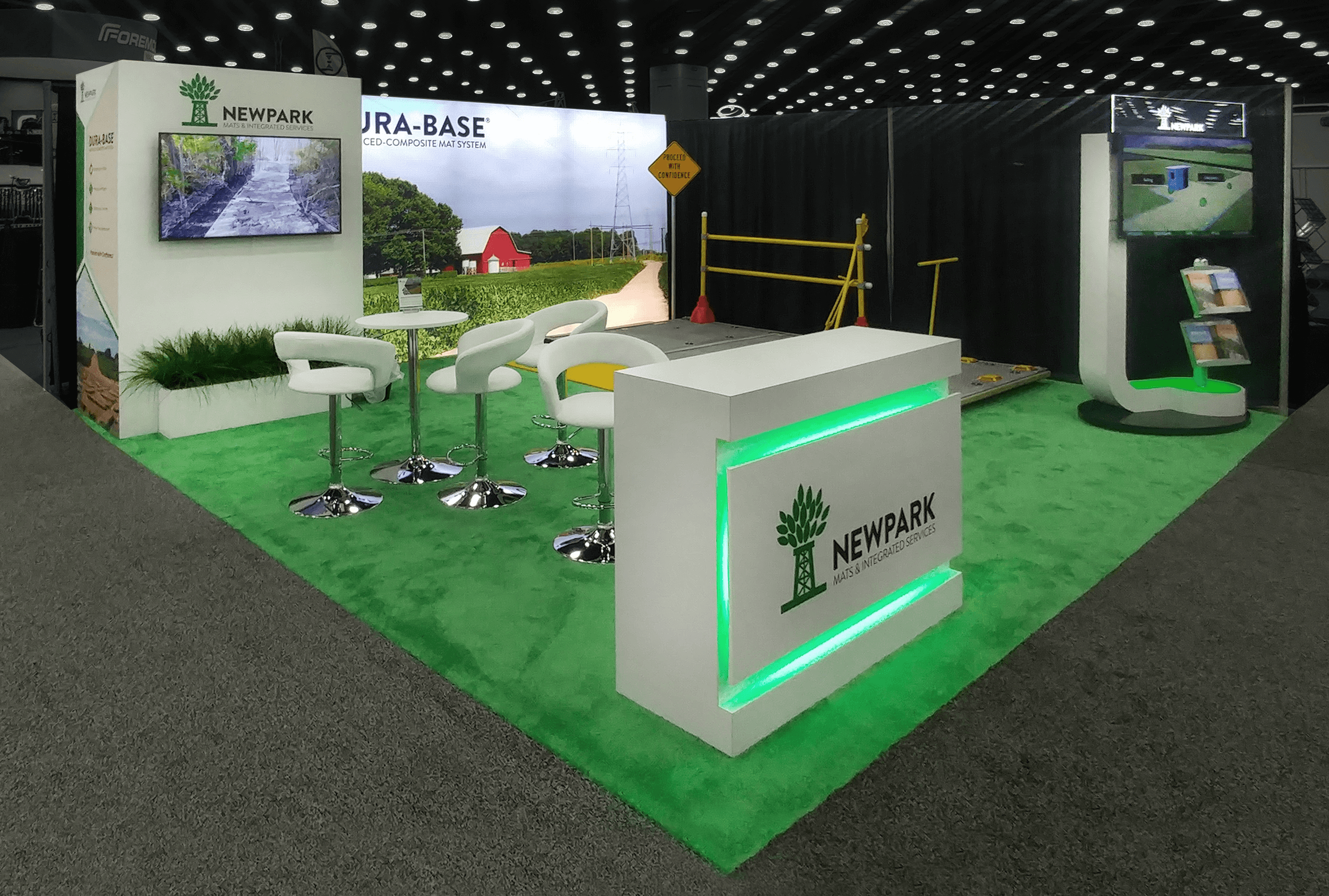 Trade show booth with green carpet, green backlit podium, white seating, a backlit graphic wall, video monitors, and games.