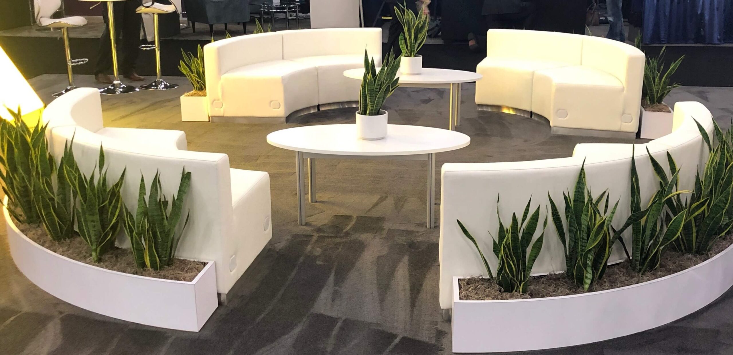 Four white, arched couches over gray padded carpet form a circular trade show seating area for customer entertainment.