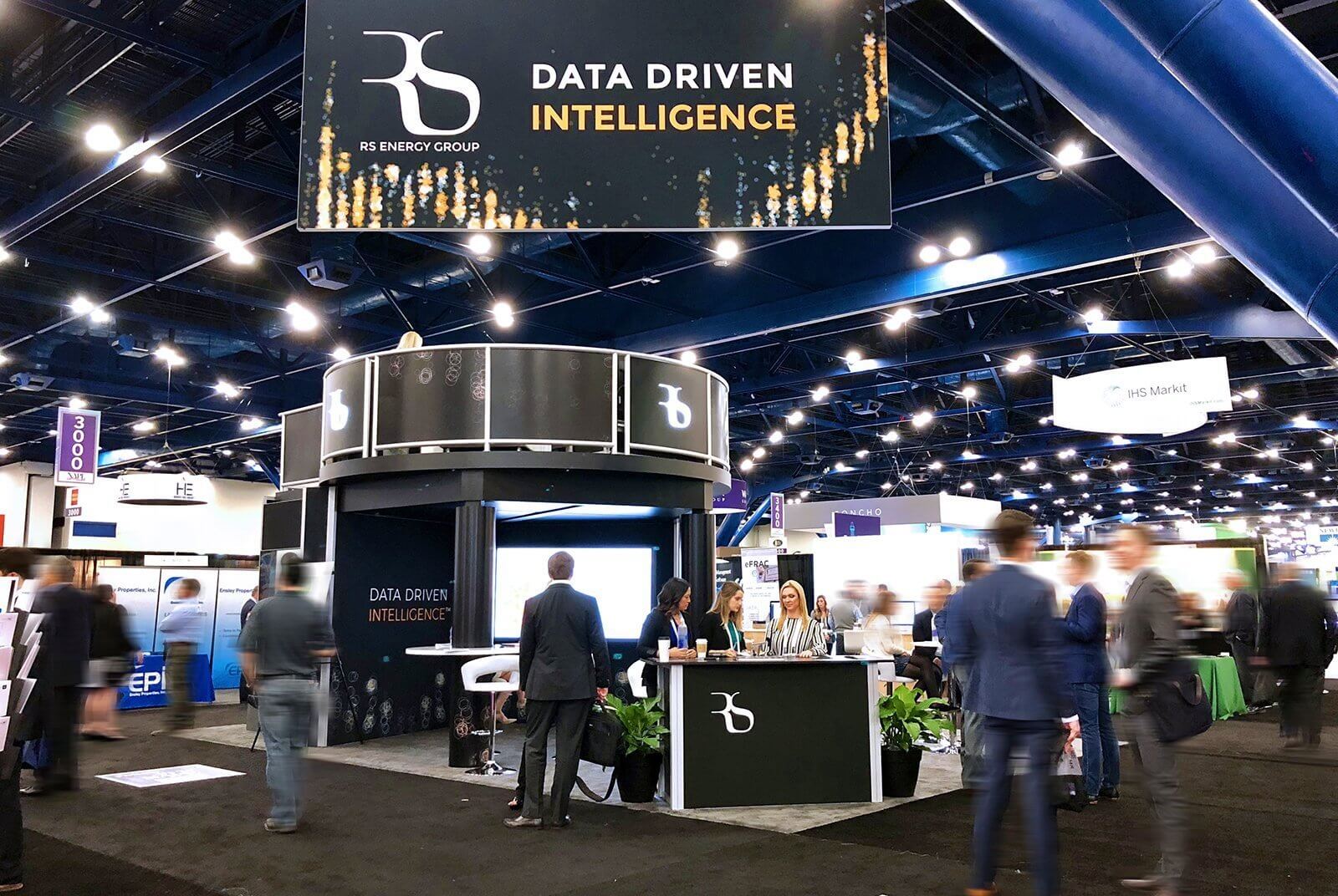Double-deck trade show booth with 3D backlit logos, seating, refrigerators, monitor, bistro tables, and a greeting counter.