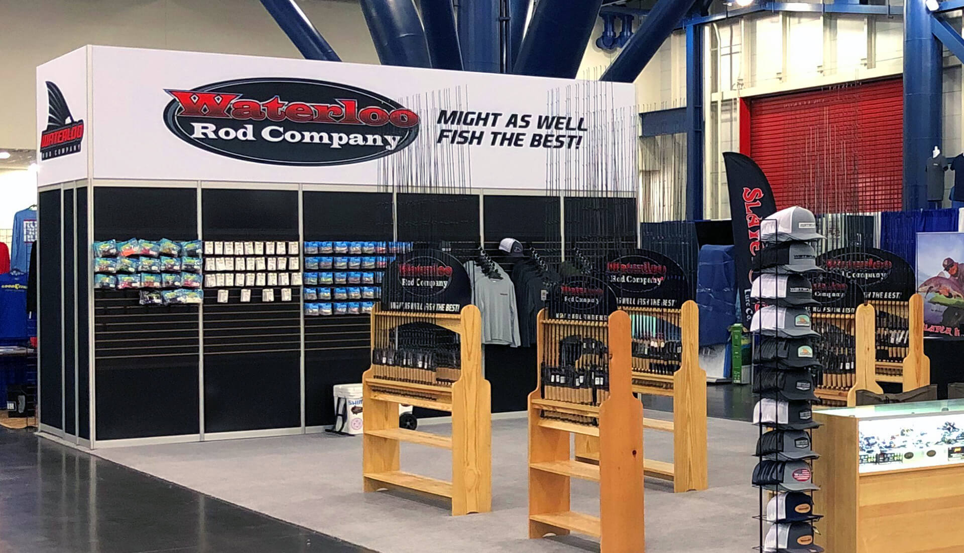 Trade show booth with lit display case, racks of fishing rods, rack of trucker hats, and back wall with hanging merchandise