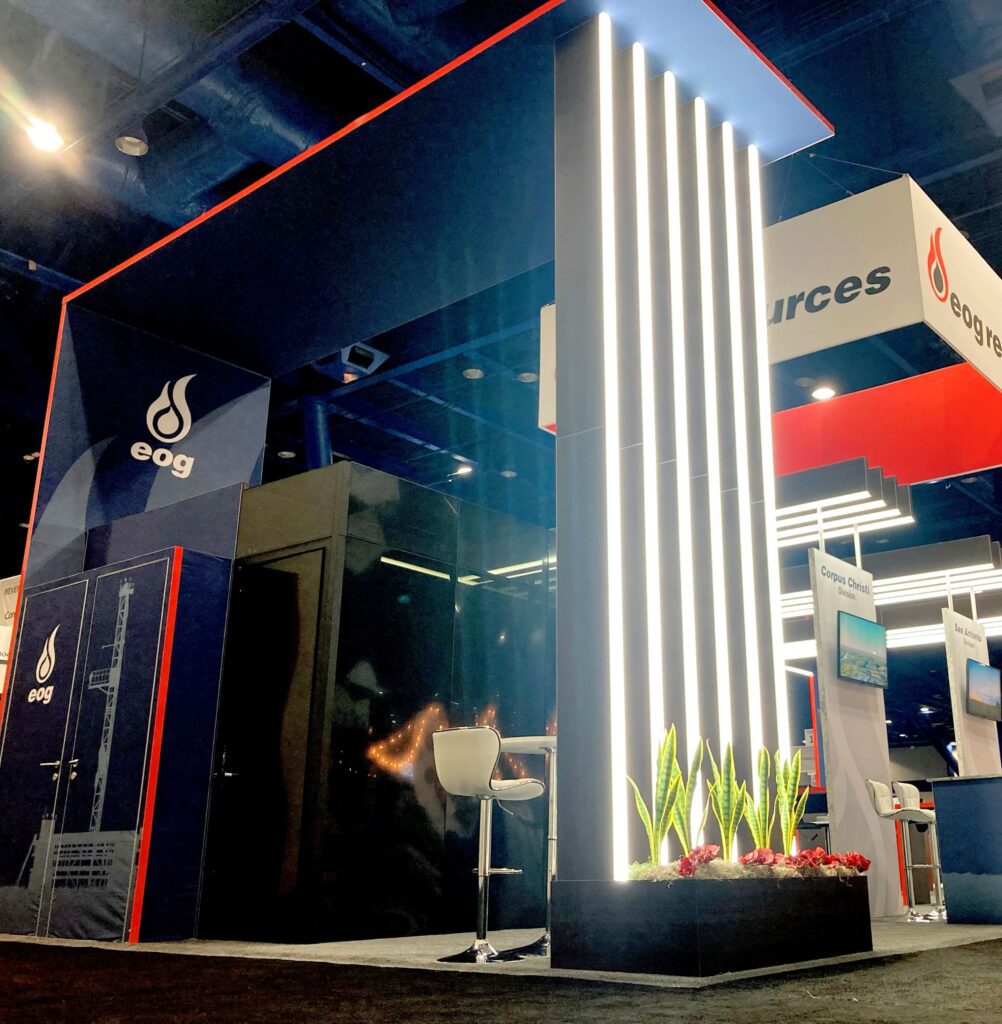 Stunning architectural booth structure with white lighted vertical pillars at each corner, large overhead branded signage, three podiums with seating, video monitors, and overhead white lighted beams